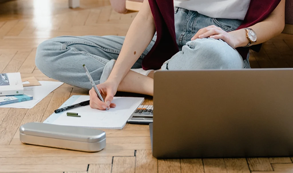 Een vrouw op de vloer met een laptop en pen, bezig met notities maken of schrijven.